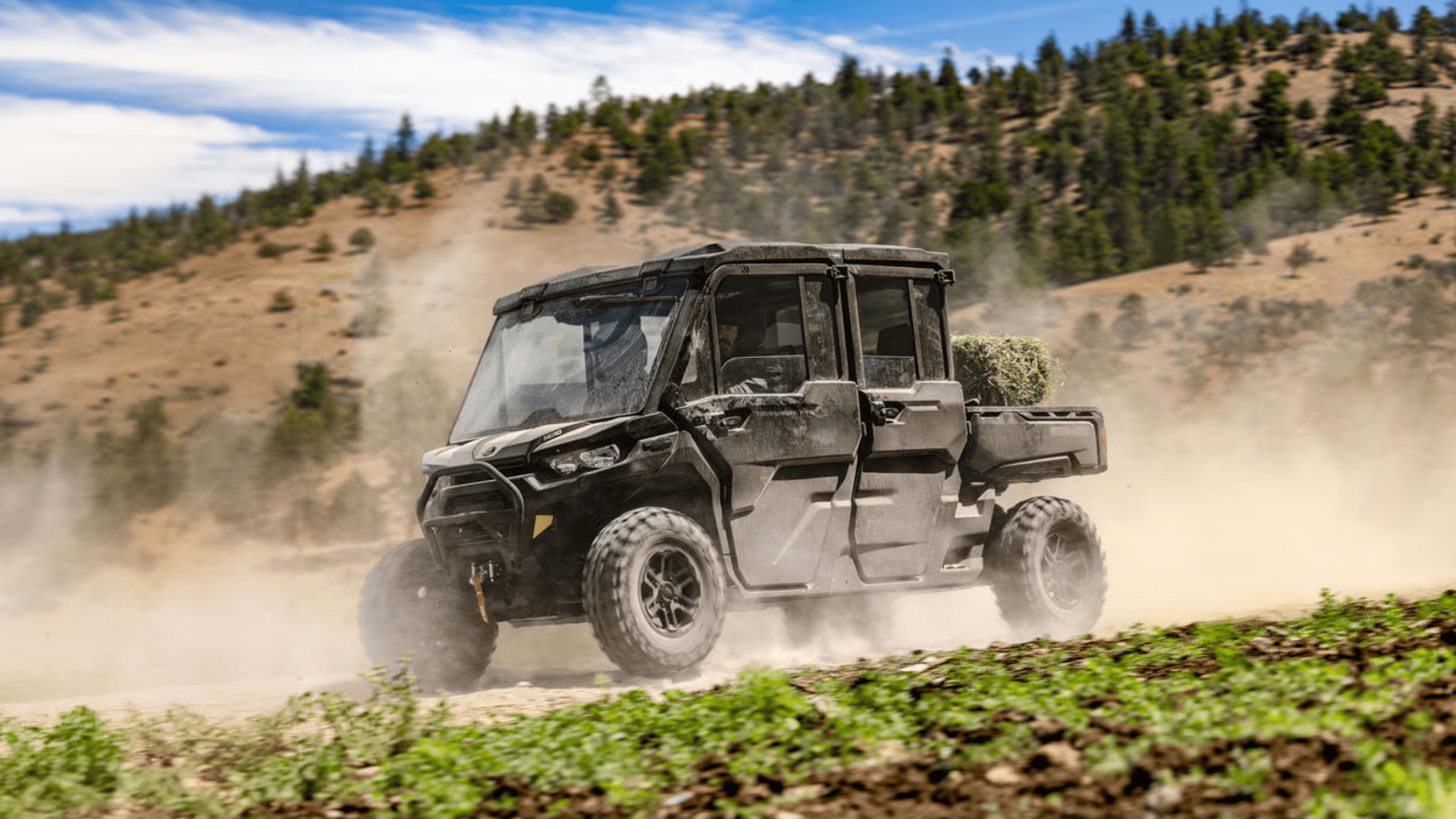 2025 Can-Am Defender hauling hay bales on a dusty trail with mountain backdrop.