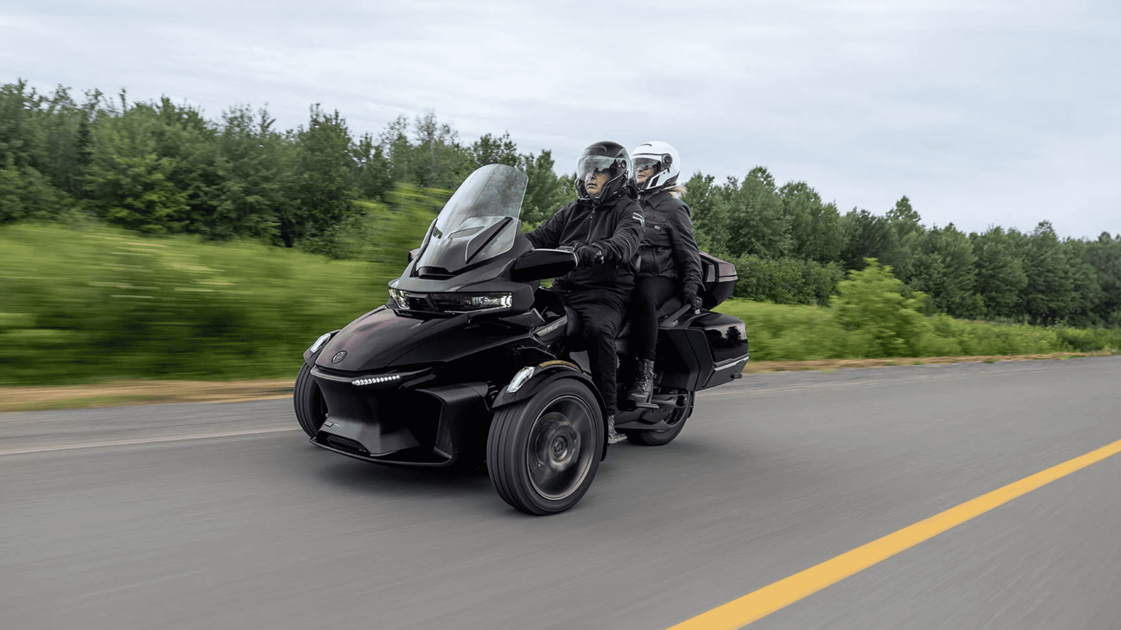 2025 Can-Am Spyder RT with two riders cruising on a highway, surrounded by greenery.