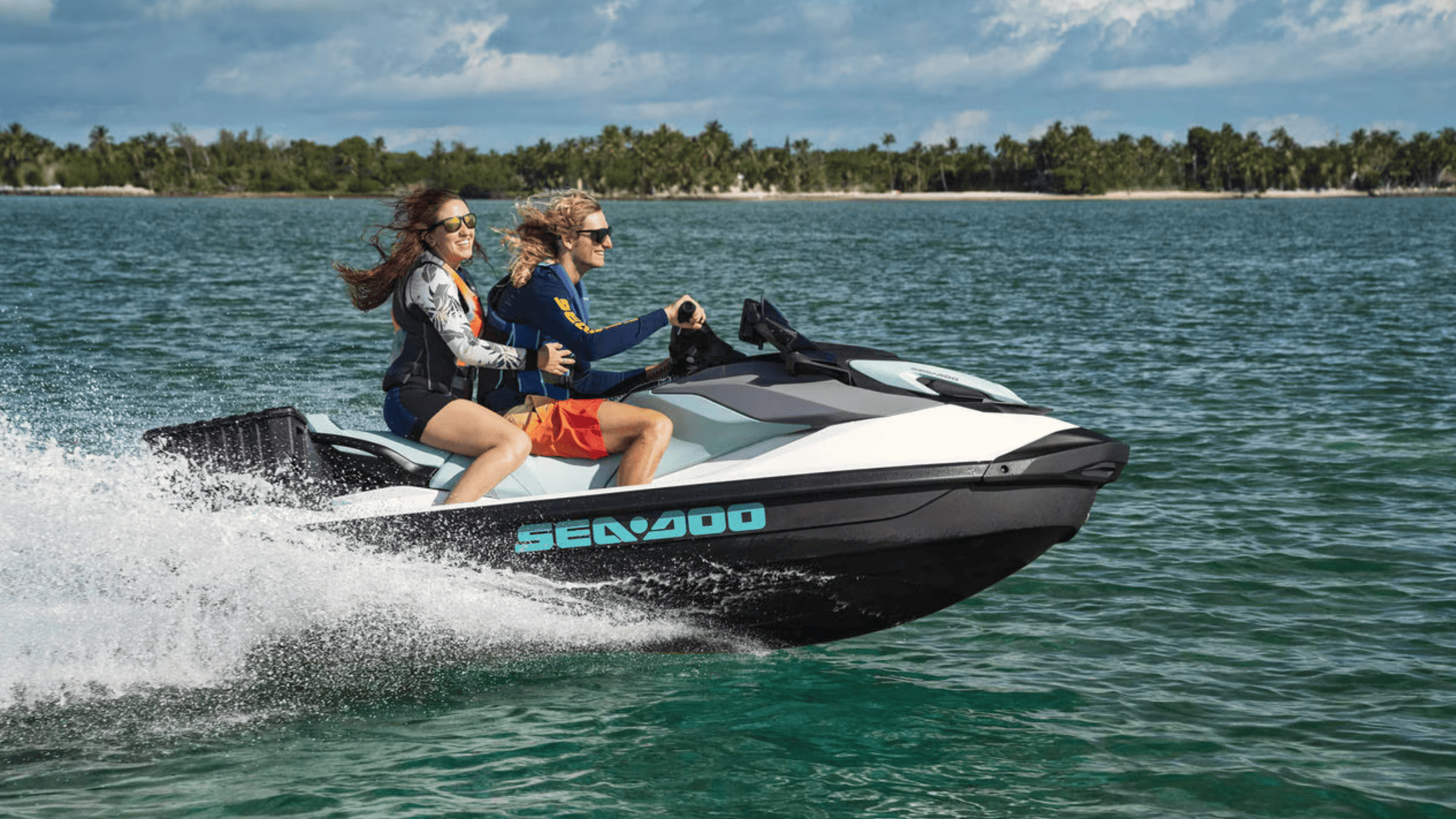 Two people riding a 2025 Sea-Doo GTI™ personal watercraft at high speed near a tropical shoreline.